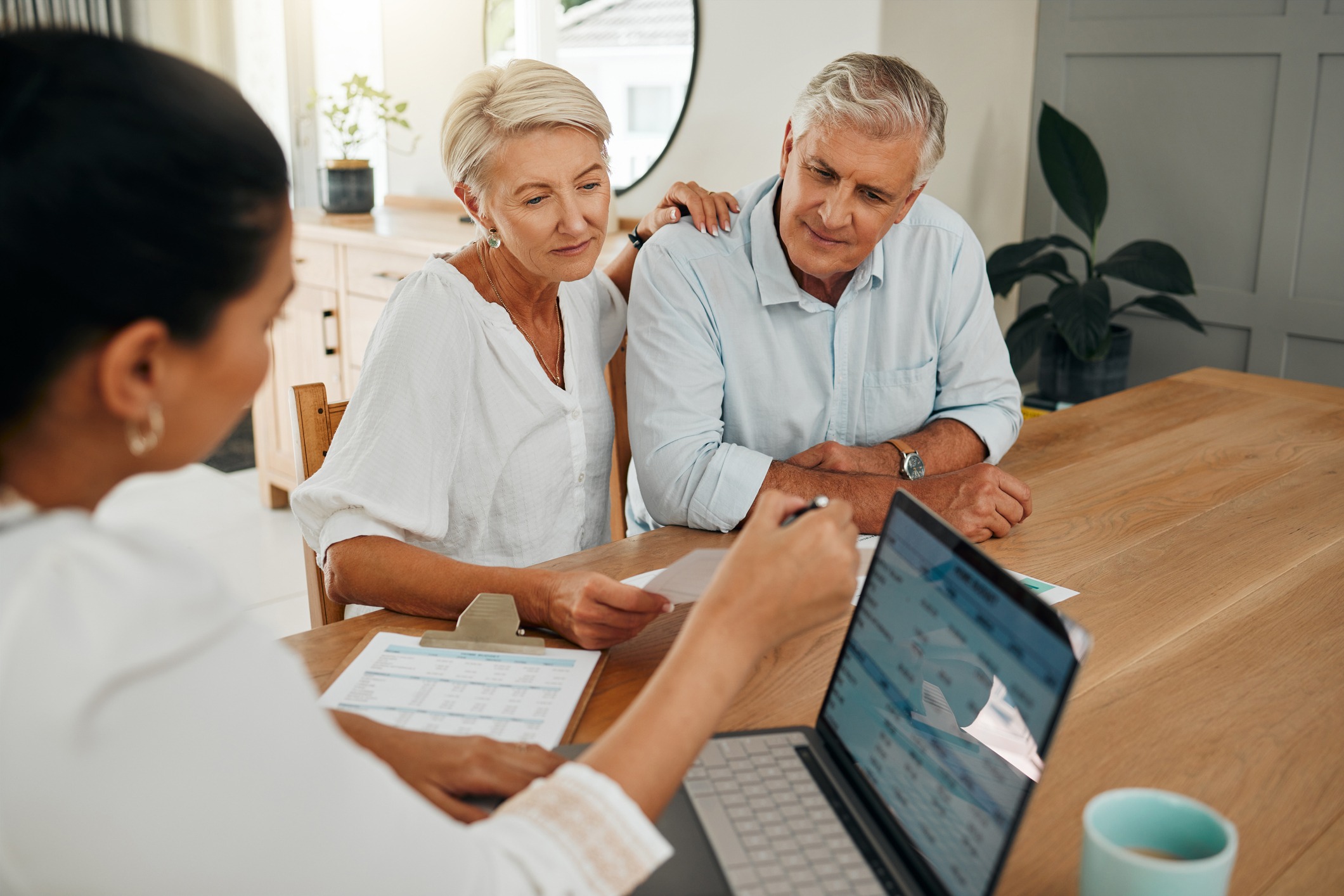 An older couple engaged in end-of-life planning at a dining room table, reviewing documents and discussing important decisions with a person using a laptop.