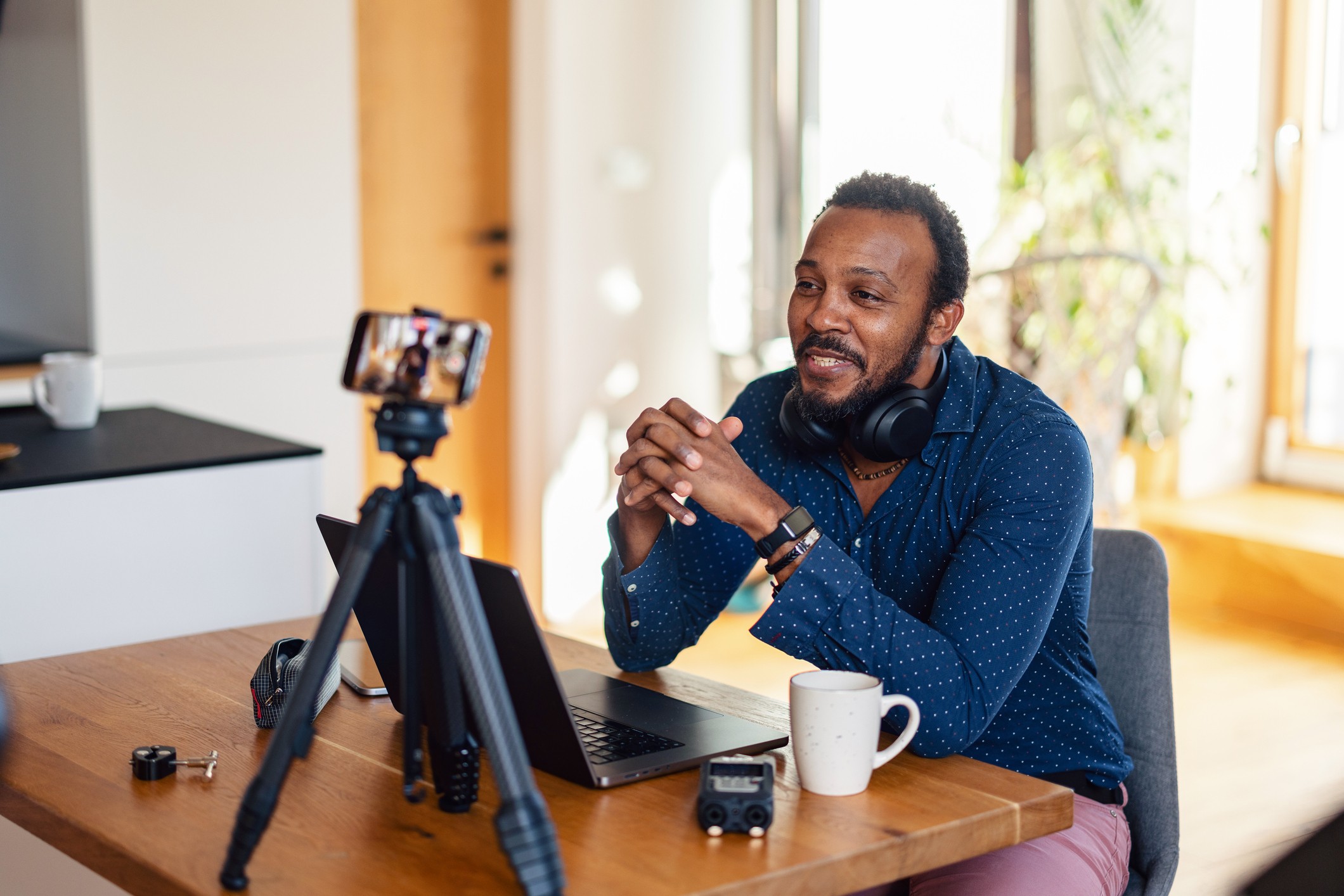 Content creator sits at a wooden table in a well-lit room, speaking about financial planning for self-employed individuals on a phone mounted on a tripod.