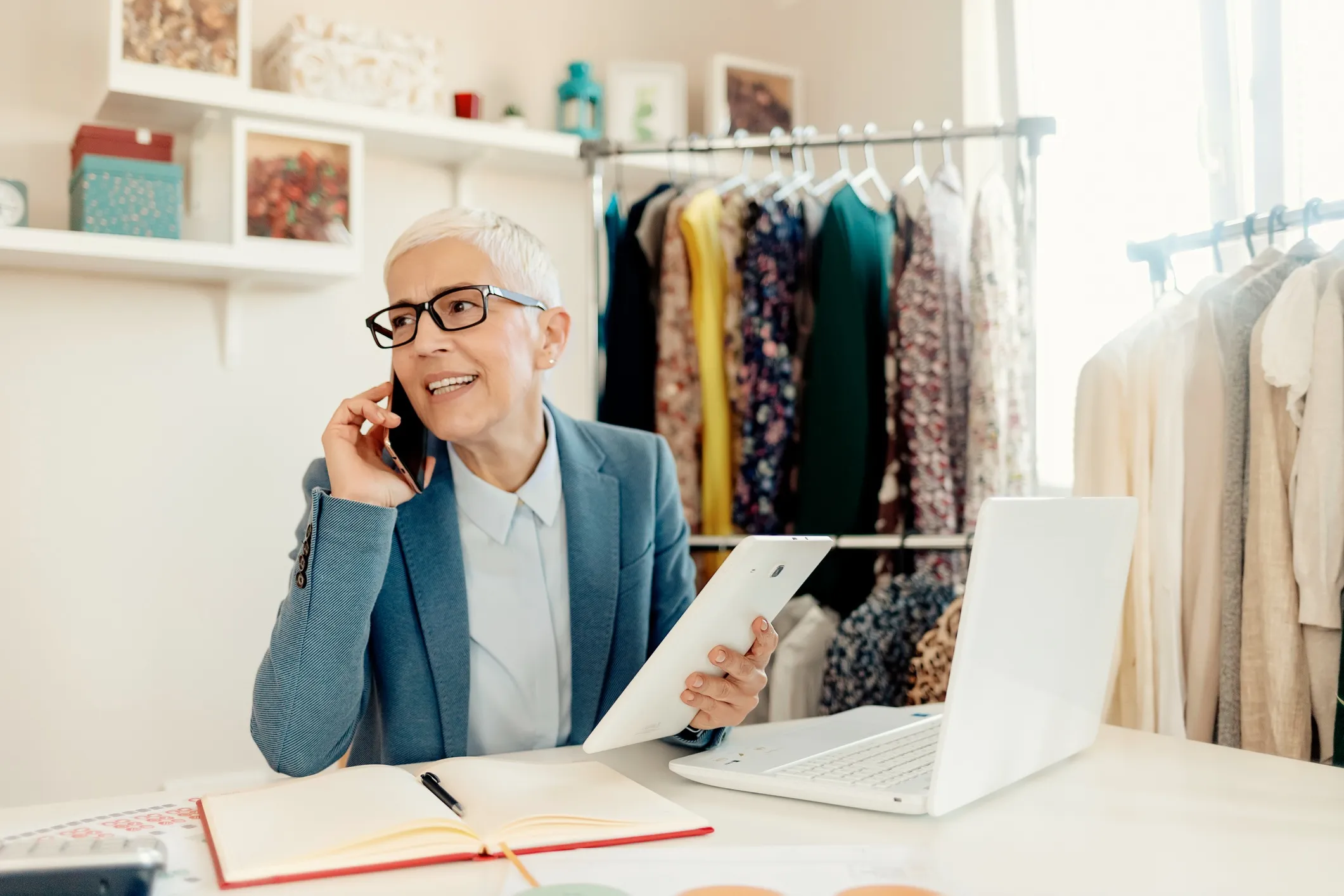 Smiling woman in a blue blazer talking on the phone about self-employed retirement plans while working at her desk with a tablet and laptop